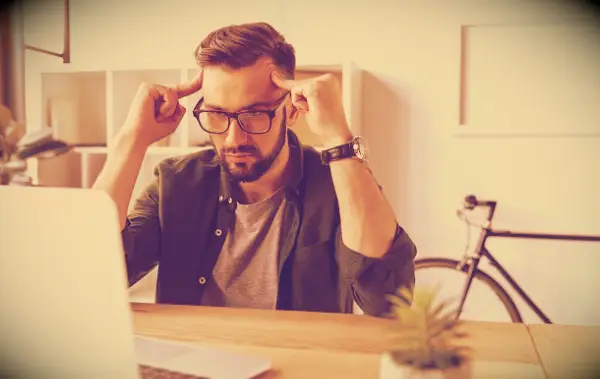A man sitting by his laptop, thinking and looking frustrated.