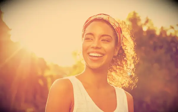 A happy and smiling woman outside with trees and the sun behind her.