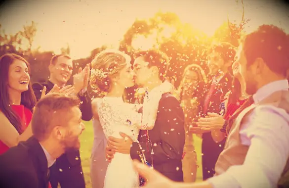 A couple kissing on their wedding day while friends around them throw confetti and cheer them on.