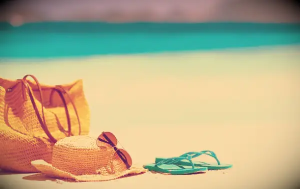 A bag, hat and flip flops in the sand at the beach with the blue water in the background.