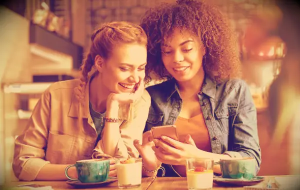 Two girls in a coffee shop laughing about something they see on their phone.