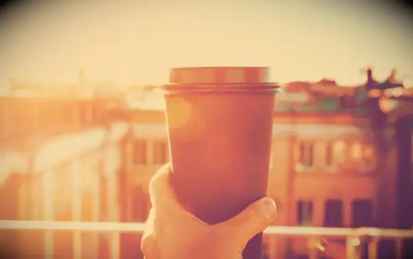 A person on a balcony with hand holding up a cup of coffee towards the sun in in the morning.